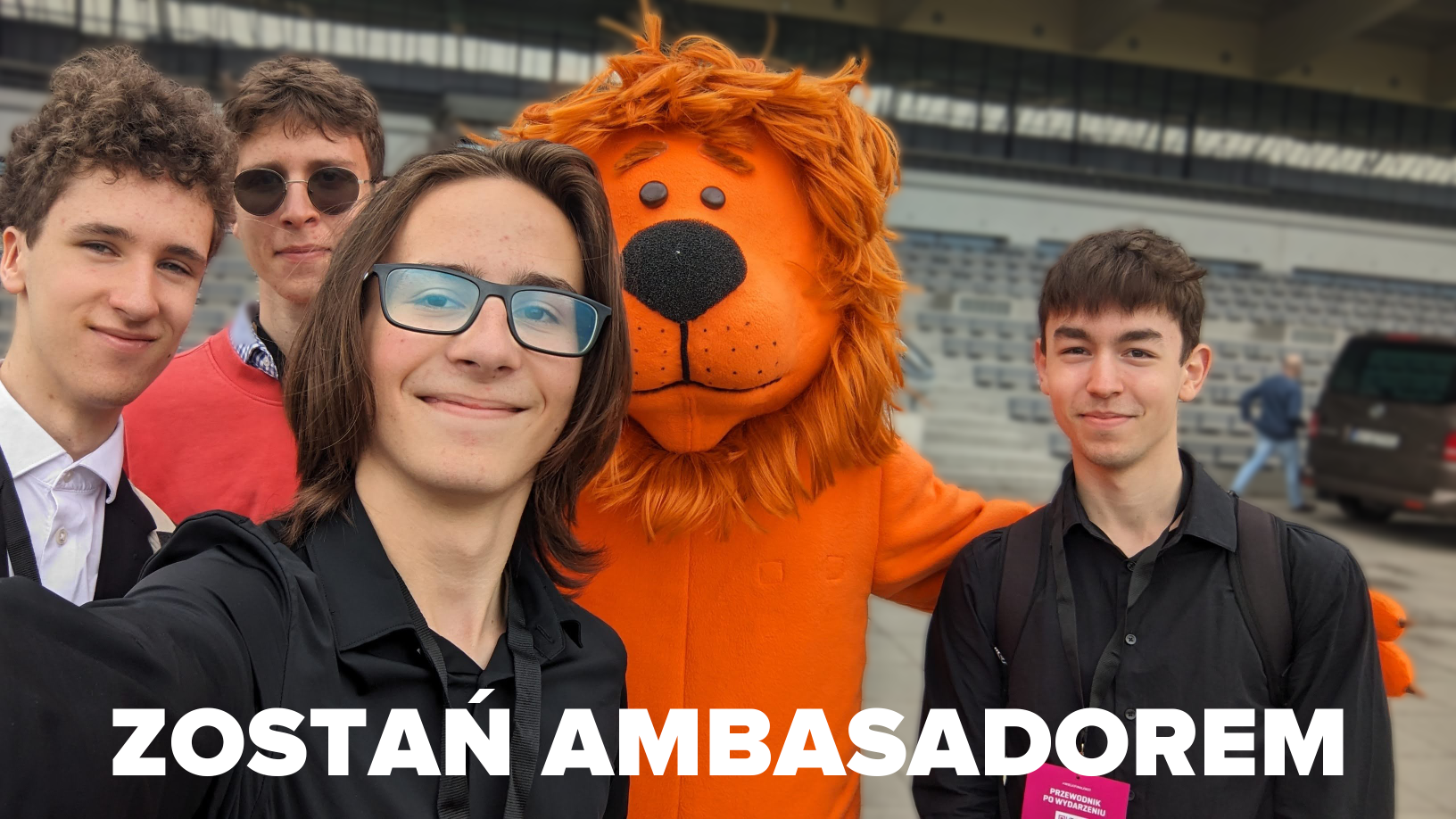 Grupa czterech młodych mężczyzn pozuje do selfie przed trybuną wraz z maskotką – dużym, pluszowym lwem o pomarańczowym futrze. Wszyscy uśmiechają się do aparatu i są ubrani w eleganckie stroje. Na zdjęciu znajduje się duży, biały napis: 'ZOSTAŃ AMBASADOREM'.